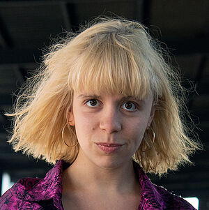 Portraitfoto von Fenja Abel. Sie läuft neben einem Gleisbett in einem Bahnhof entlang. Ihre Arme sind verschränkt. Sie trägt eine lilafarbene gemusterte Bluse zu einer hellen Jenas. Sie hat blondes schulterlanges Haar mit Pony.