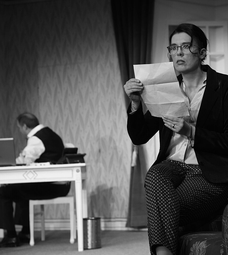 Schwarz-weißes Foto. Barbara Fürcht sitzt auf der Armlehne eines Sessels und hält einen Brief in den Händen. Sie schaut in die Luft und wirkt wie eingefroren. Im Hintergrund ist Dr. Richard Fürcht verschwommen zu sehen. Er sitzt auf einem Stuhl an einem Schreibtisch und ist von Barbara abgewandt. 