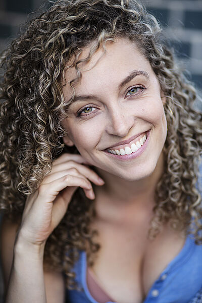 Portraitfoto von Nicole Rushing. Ihr Kopf lehnt auf ihrer rechten Hand, der Kopf ist leicht geneigt. Sie grinst mit offenem Mund. Ihre feinen Locken fallen ihr auf die Schultern. Sie trägt ein hellblaues Top. Im Hintergrund ist eine Ziegelmauer zu erahnen.