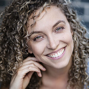 Portraitfoto von Nicole Rushing. Ihr Kopf lehnt auf ihrer rechten Hand, der Kopf ist leicht geneigt. Sie grinst mit offenem Mund. Ihre feinen Locken fallen ihr auf die Schultern. Sie trägt ein hellblaues Top. Im Hintergrund ist eine Ziegelmauer zu erahnen.