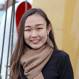 Portraitfoto von Nozomi Nambara. Sie lächelt mit geöffnetem Mund. Sie trägt einen beigefarbenen Schal und ein dunkles Oberteil. Ihr dunkelbraunes glattes Haar fällt uhr auf die Schultern. Sie trägt goldene Ohrringe.