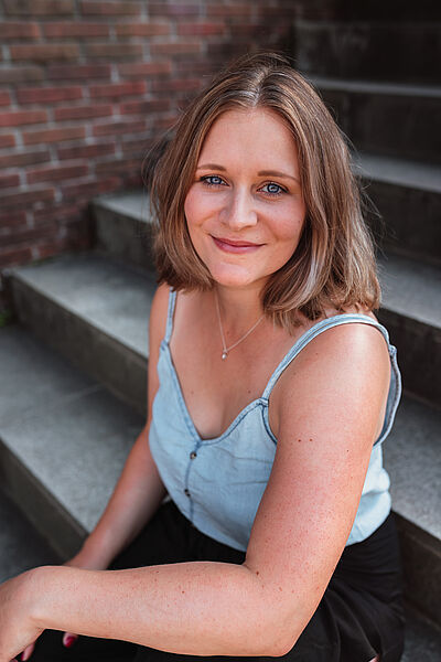 Portraitfoto von Nikola Frehsee. Sie sitzt auf einer Treppe. Ihr linker Arm ist auf ihrem Schoß aufgelegt. Sie blickt lächelnd nach oben. Sie trägt schulterlanges blondes Haar, ein hellblaues Jeans-Top und eine schwarze Stoffhose.