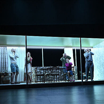 Mehrere Menschen stehen im Haus. Es wird blau beleuchtet. Sie halten sich die Köpfe fest und schreien.