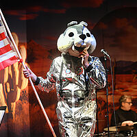 Ein silber gekleideter Mensch mit einem Nagerkostüm auf dem Kopf und der Flagge der USA in der rechten Hand. Im Hintergrund spielt ein Schlagzeuger. 