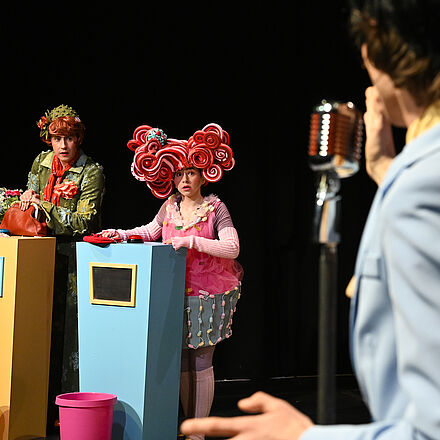 Im Vordergrund steht ein Mann mit dem Rücken zur Kamera vor einem Mikrofon. Im Hintergrund stehen zwei Personen hinter jeweils einem Pult mit einem Knopf oben drauf.