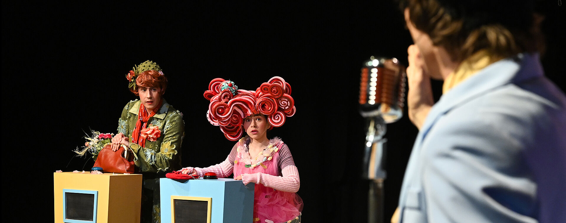 Im Vordergrund steht ein Mann mit dem Rücken zur Kamera vor einem Mikrofon. Im Hintergrund stehen zwei Personen hinter jeweils einem Pult mit einem Knopf oben drauf.