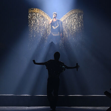 Im Hintergrund steht ein Mann mit goldenen Flügeln auf einer Erhöhung. Im dunklen Vordergrund tanzt eine Frau im Schatten.