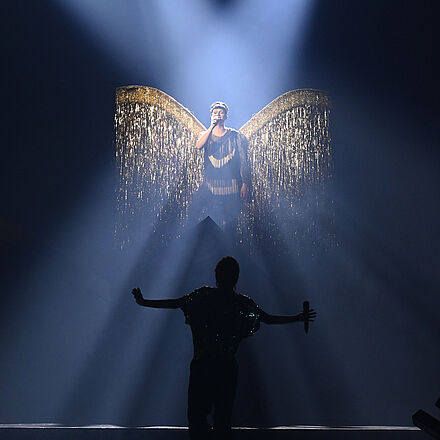 Im Hintergrund steht ein Mann mit goldenen Flügeln auf einer Erhöhung. Im dunklen Vordergrund tanzt eine Frau im Schatten.