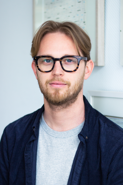 Portraitfoto von Rennik-Jan Neggers. Er hast dunkelblondes Haar, welches er zum Mittelscheitel trägt und ihm zu beiden Seiten auf die Stirn fällt. Er trägt eine auffällige dunkle Brille mit rechteckigen Gläsern. Außerdem trägt er einen Bart. Er ist in ein offenes dunkelblaues Hemd gekleidet, darunter trägt er ein hellgraues T-Shirt. Im Hintergrund ist eine weiße Wand mit einem Bilderrahmen.