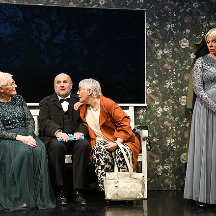 Zwei Frauen in Abendkleidung und ein Mann sitzen auf der Bank. Eine Frau steht im Abendkleid daneben.