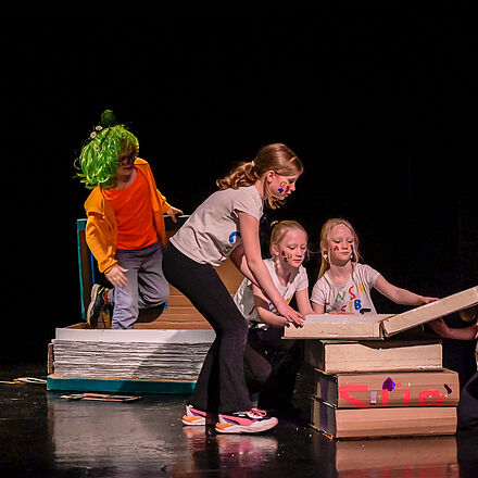 Vier Mädchen schlagen ein überdimensioniertes Buch auf. Sie tragen weiße T-Shirts mit bunten Buchstaben. Auf ihren Wangen sind bunte Buchstaben gemalt. Im Hintergrund steigt ein weiteres Kind mit grüner Perücke aus einem aufgeschlagenen Buch heraus.