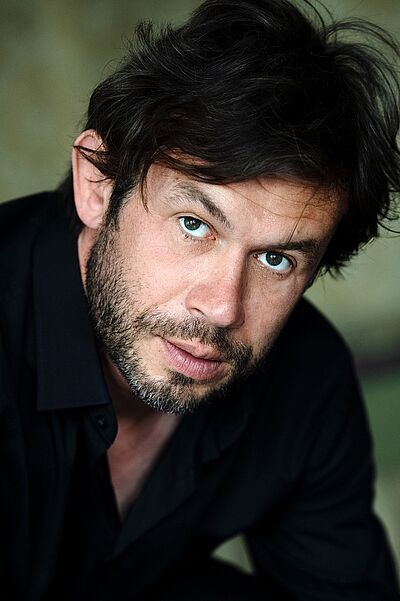 Portraitfoto von Martin Maecker. Er sitzt nach vorn gebeugt. Er hat dunkles Haar, dass ihm locker über den Kopf fällt, dazu ein Dreitagebart. Er trägt ein schwarzes Hemd, dessen oberer Knopf geöffnet ist.