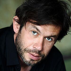 Portraitfoto von Martin Maecker. Er sitzt nach vorn gebeugt. Er hat dunkles Haar, dass ihm locker über den Kopf fällt, dazu ein Dreitagebart. Er trägt ein schwarzes Hemd, dessen oberer Knopf geöffnet ist.