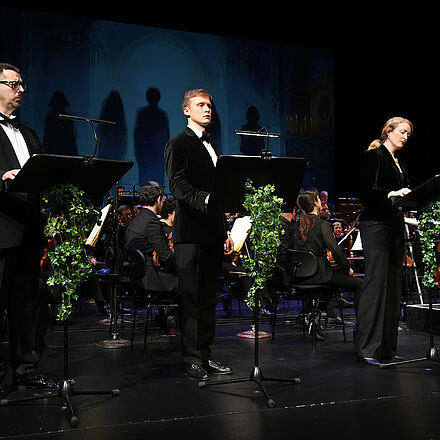 Im Vordergrund stehen drei Personen und singen, im Hintergrund ist das Orchester erkennbar.  