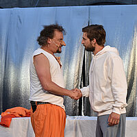 Zwei Cyrano und Christian stehen sich händeschüttelnd gegenüber und blicken sich an. Der Schauspielende im orangenen Anzug hat sein Jackett ausgezogen und trägt eine große Nasenprothese. Im Hintergrund glänzt ein silbernes Tuch als Teil des Bühnenbilds.