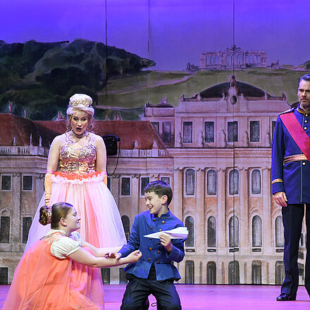 Im Hintergrund ist ein Schloss zu sehen. Zwei Erwachsene stehen vor zwei spielenden Kindern