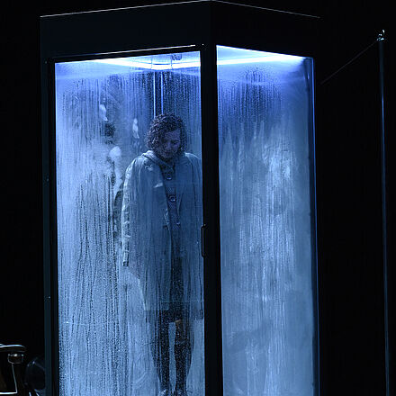 Eine Frau steht in einer Box. Sie schaut auf den Boden. Es regnet.