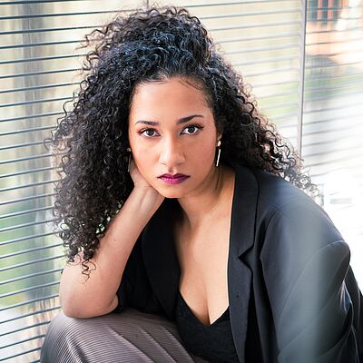 Portraitfoto von Jamie-Lee Uzoh. Ihr Kopf lehnt auf ihrer rechten Hand. Der Ellenbogen des rechten Arms ist auf ihrem hochgelegten rechten Bein abgelegt. Ihr schwarzes lockiges Haar fällt ihr über die rechte Schulter. Sie trägt einen schwarzen Blazer und ein schwarzes Oberteil. Im Hintergrund sind geschlossene Jalousien zu sehen.