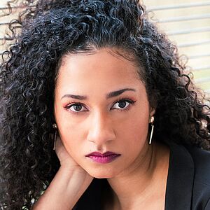 Portraitfoto von Jamie-Lee Uzoh. Ihr Kopf lehnt auf ihrer rechten Hand. Der Ellenbogen des rechten Arms ist auf ihrem hochgelegten rechten Bein abgelegt. Ihr schwarzes lockiges Haar fällt ihr über die rechte Schulter. Sie trägt einen schwarzen Blazer und ein schwarzes Oberteil. Im Hintergrund sind geschlossene Jalousien zu sehen.