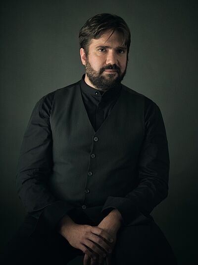 Portraitfoto von Davide Cabassi. Er ist schwarz gekleidet und sitzt vor einem schwarzen indirekt beleuchtetem Hintergrund. Er trägt eine Weste über einem Hemd. Seine Hand sind unter dem Bauch übereinandergelegt. Er trägt eine schwarze Kurzhaarfrisur und einen Vollbart im Gesicht.