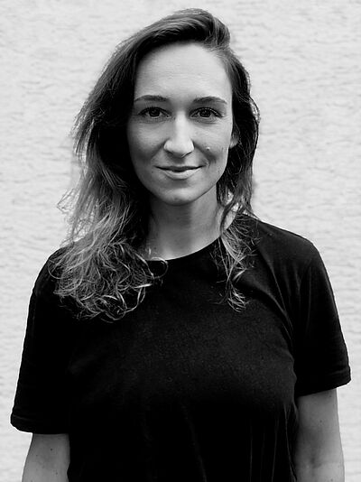 Schwarzweißes Portraitfoto von Ariane Königshof. Sie steht in einem schwarzen T-Shirt vor einer weißen Wand. Ihr lockiges Haar hängt ihr auf die Schultern. Sie lächelt leicht.