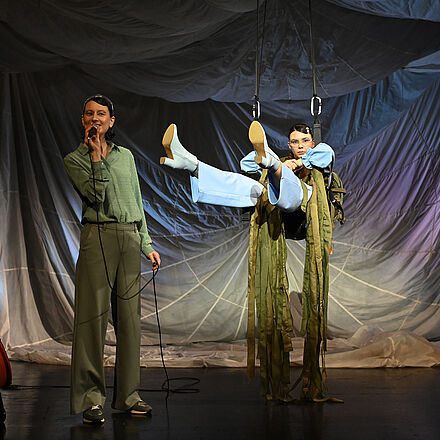 Eine Frau steht am Mikrofon. Eine andere Frau hängt von der Decke und schwingt auf einer Schaukel.