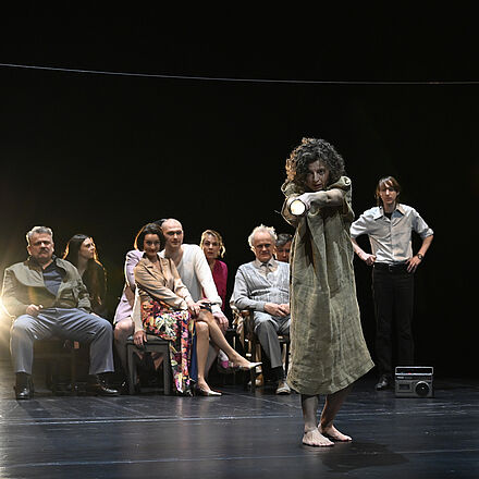 Eine Frau hält eine Taschenlampe in die Kamera. Im Hintergrund sitzen Personen und schauen ihr dabei zu.