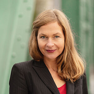 Portraitfoto von Sibylla Rasmussen. Ihr Haar hängt auf die Schultern herab, eine Strähne ist mit einer Haarklemme neben dem Gesicht festgesteckt. Sie trägt einen schwarzen geschlossenen Blazer, darunter ein rotes Oberteil. Im Hintergrund sind verschwommen grüne Stahlstreben zu sehen.