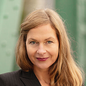 Portraitfoto von Sibylla Rasmussen. Ihr Haar hängt auf die Schultern herab, eine Strähne ist mit einer Haarklemme neben dem Gesicht festgesteckt. Sie trägt einen schwarzen geschlossenen Blazer, darunter ein rotes Oberteil. Im Hintergrund sind verschwommen grüne Stahlstreben zu sehen.