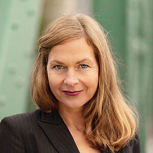 Portraitfoto von Sibylla Rasmussen. Ihr Haar hängt auf die Schultern herab, eine Strähne ist mit einer Haarklemme neben dem Gesicht festgesteckt. Sie trägt einen schwarzen geschlossenen Blazer, darunter ein rotes Oberteil. Im Hintergrund sind verschwommen grüne Stahlstreben zu sehen.