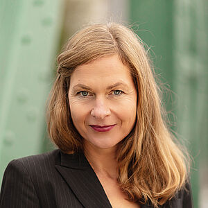 Portraitfoto von Sibylla Rasmussen. Ihr Haar hängt auf die Schultern herab, eine Strähne ist mit einer Haarklemme neben dem Gesicht festgesteckt. Sie trägt einen schwarzen geschlossenen Blazer, darunter ein rotes Oberteil. Im Hintergrund sind verschwommen grüne Stahlstreben zu sehen.