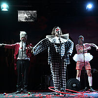 Drei Sänger:innen in Clownskostümen. Um sie herum liegen Bälle, Gewichte und ein Hulahoop-Reifen. 