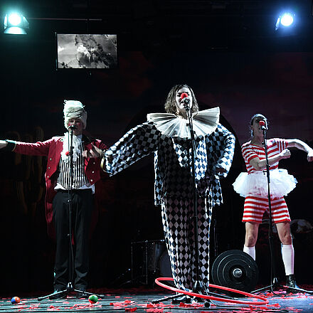 Drei Sänger:innen in Clownskostümen. Um sie herum liegen Bälle, Gewichte und ein Hulahoop-Reifen. 