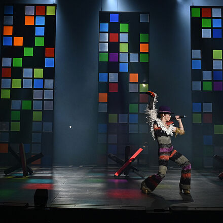 Im Hintergrund stehen drei bunte Glaswände. Auf dem Boden befinden sich mehrere Grenzblockaden. Im Vordergrund steht eine Frau, die in der einen Hand ein Mikrofon hält und den anderen Arm nach oben streckt.