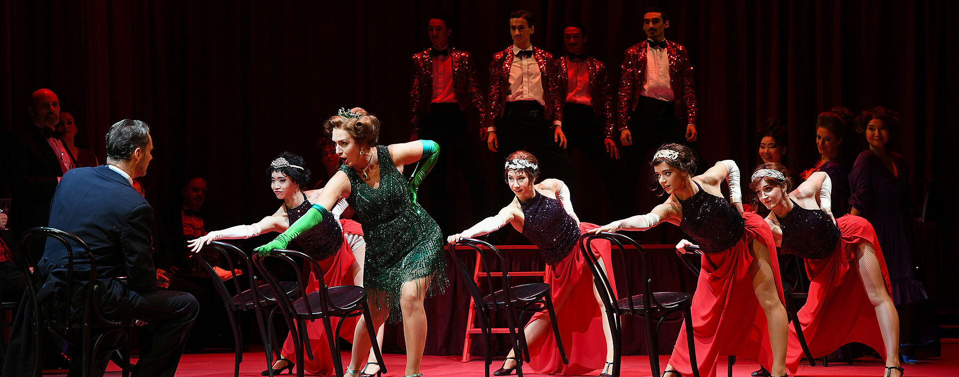 Im Hintergrund stehen Männer in glitzernden Anzügen auf der Bühne. Davor tanzen Frauen an Stühlen und blicken auf einen Mann.