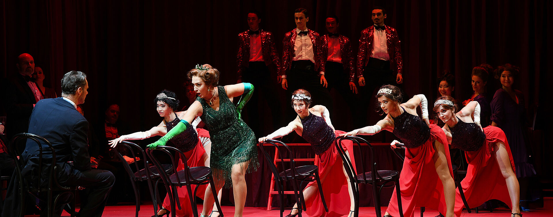 Im Hintergrund stehen Männer in glitzernden Anzügen auf der Bühne. Davor tanzen Frauen an Stühlen und blicken auf einen Mann.