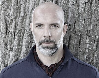 Portraitfoto von Bart Driessen. Er lehnt an einem Baum. Er trägt ein dunkelblaues Oberteil, dessen Reißverschluss oben leicht geöffnet ist. Darunter ist ein dunkles Tuch. Sein Kopf ist kahl. Er trägt einen gräulich-weißen gepflegten Vollbart.