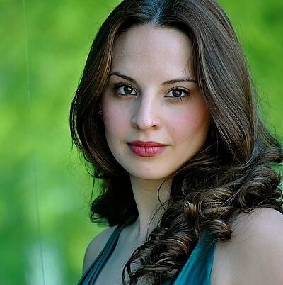 Portraitfoto von Sydney Gabbard. Sie blickt seitlich gedreht in die Kamera. Ihr braunes Haar ist zu Wellen geformt und fallen ihr über die Schulter. Sie trägt ein dunkeltürkises Kleid. Der Hintergrund ist grün und verschwommen.