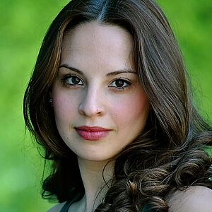 Portraitfoto von Sydney Gabbard. Sie blickt seitlich gedreht in die Kamera. Ihr braunes Haar ist zu Wellen geformt und fallen ihr über die Schulter. Sie trägt ein dunkeltürkises Kleid. Der Hintergrund ist grün und verschwommen.