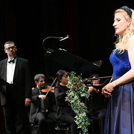 Im Vordergrund steht eine Person in einem blauen Kleid, ein Mann schaut sie an. Im Hintergrund spielt das Orchester.  