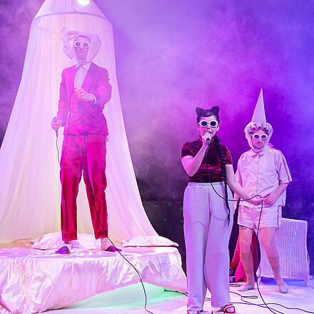Drei Personen mit weißen Sonnenbrillen performen einen Song. Die Bühne ist in Nebel gehüllt der in violetten Farbtönen schimmert.