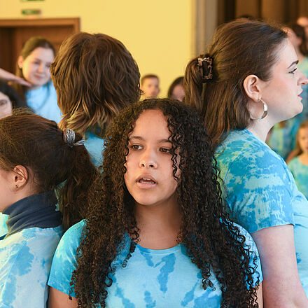 Im Mittelpunkt des Bildes stehen vier Mädchen. Im Hintergrund steht der restliche Kinderchor. Alle tragen ein blaues Oberteil.