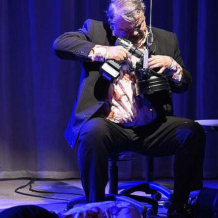 Im unteren Vordergrund liegt eine Frau auf dem Boden. Im Mittelpunkt des Bildes sitzt ein Mann auf einem Stuhl und hält einen Akkuschrauber an eine Lampe.