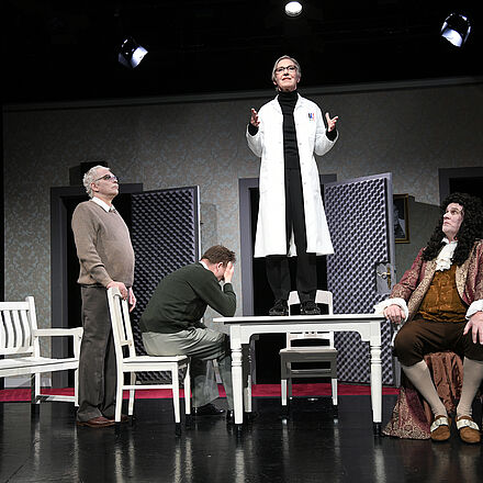 Im Mittelpunkt des Bildes steht eine Frau mit Arztkittel auf einem Tisch. Um den Tisch herum stehen drei Männer. Der rechte Mann trägt Kleidung aus der Barock-Zeit.