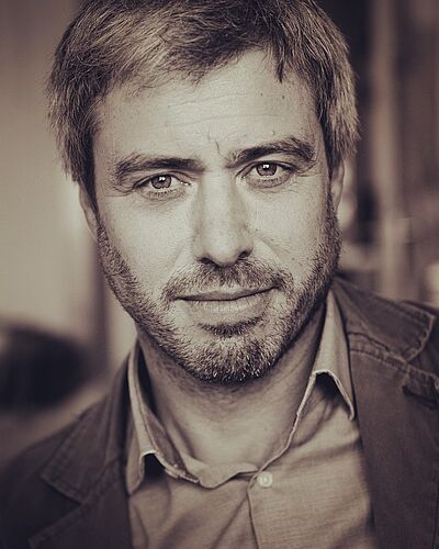Sepiafarbenes Portraitfoto von Sebastian Hölz. Er hat kurzes Haar, trägt einen Vollbart, ein helles Hemd und darüber eine Jacke.