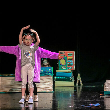 Zwei Kinder tanzen auf einer Bühne. Um sie herum liegen überdimensionierte Bücher. Aus einem schaut ein Junge heraus.