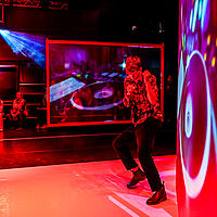 Lenny tanzt. Er steht zwischen Leinwänden auf die ein DJ-Pult projiziert wird. Die Szenerie ist in rotes Licht gehüllt. Im Hintergrund sitzt das Publikum, über ihnen steht ein Ray an einem DJ-Pult und hält ein Mikrofon in den Händen,.