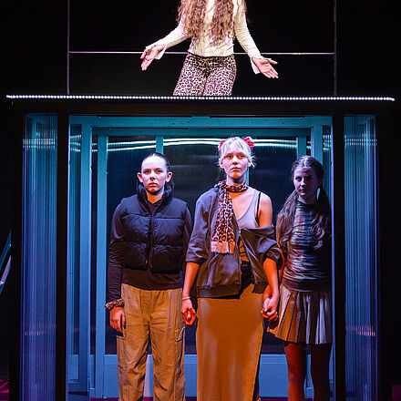 Drei der Mädchen stehen im Gebäude, ernster Blick und halten sich an den Händen fest. Auf dem Dach kniet ein weiteres Mädchen. 