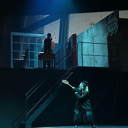 Eine Frau mit einem Teppichklopfer steht im Vordergrund. Man erkennt im Hintergrund einen Mann und eine Frau in weiß.