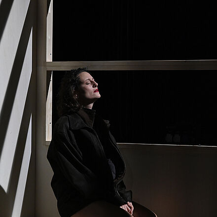 Die Frau sitzt am Fenster mit geschlossenen Augen. Sie sitzt zwischen den Federn, das Licht fällt auf ihr Gesicht.